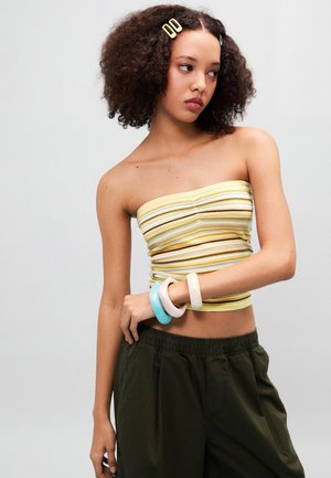 Jeune femme aux cheveux bouclés portant un top bandeau jaune à rayures, un pantalon sombre et des bracelets colorés, regardant de côté avec une expression neutre.