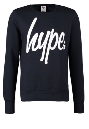 Mężczyzna idący, ubrany w czarną bluzę "hype.", szare jeansy, czarne trampki, niosący okrągłą beżową torbę z brązowymi uchwytami.