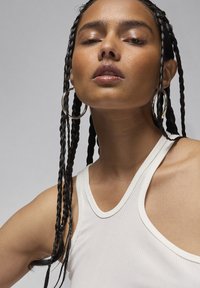 Top in cotone bianco a coste con scollo rotondo. Il modello ha i capelli lunghi e intrecciati e indossa grandi orecchini a cerchio in argento. Texture della pelle liscia.