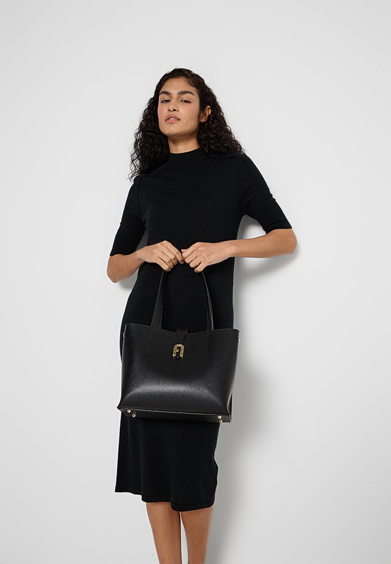 Donna in un vestito nero che tiene una grande borsa in pelle nera con chiusura dorata, in piedi contro uno sfondo grigio chiaro uniforme.