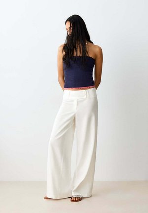 Mujer de pie junto a una pared blanca, que lleva un top azul marino sin tirantes con textura y ribete rosa, y pantalones blancos de pierna ancha con sandalias.