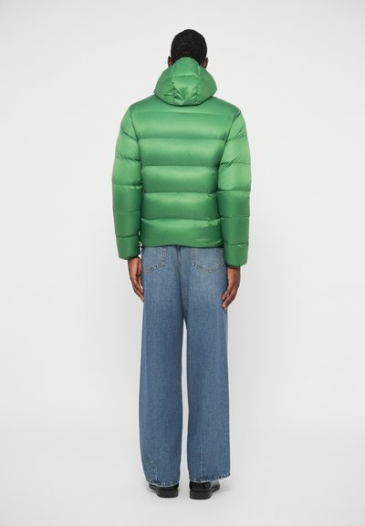 Zöld puffdzseki vízszintes tűzéssel, kapucnival és rugalmas mandzsettaakkal, széles kék farmernadrággal és fekete cipővel párosítva.