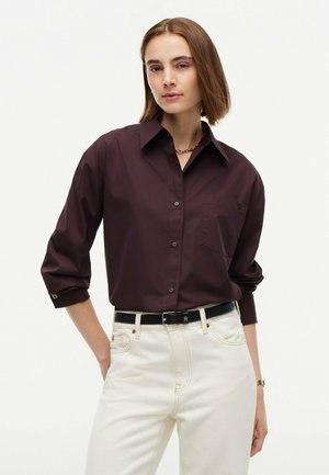 Woman with short brown hair wearing a dark purple button-up shirt with rolled sleeves and white high-waisted pants with a black belt.