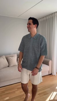 Īsām piedurknēm, strukturēts pelēks adīts krekls ar sarežģītiem rakstiem un pogu aizdari, apvienots ar vieglām, platām baltām šortiem.