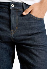 Dunkelblaue Jeans aus Denim mit oranger Naht, einer Vorderreißverschlusstasche und einem Metallknopfverschluss. Die Stoffoberfläche wirkt glatt mit einem leicht abgenutzten Look.