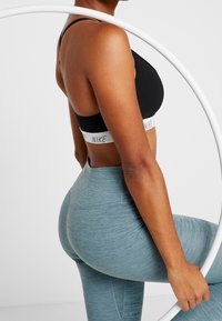 Donna in reggiseno sportivo nero Nike e leggings blu, con un hula hoop bianco in mano, in piedi di fronte a uno sfondo bianco uniforme.