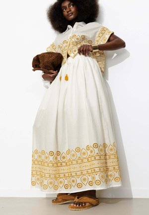 Femme aux cheveux naturels portant une robe blanche avec une broderie florale jaune, tenant un sac marron imprimé léopard et portant des sandales marron.