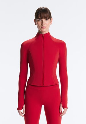 Žena oblečená v priliehavom červenom bundovom tričku na zips s dlhým rukávom a zodpovedajúcich červených legínach, stojaca na jednoduchom svetlom pozadí.