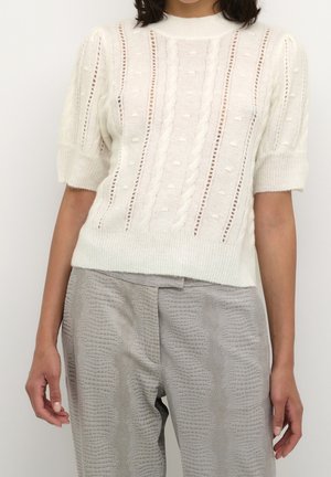 Nainen pukeutuneena valkoiseen lyhythihaiseen neulepuseroon, jossa on kaapelikuviot, ja vaaleanharmaisiin teksturoiduihin housuihin, seisoo yksiväristä taustaa vasten.