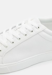 White sneaker featuring a textured perforated pattern, flat white laces, and metal eyelets. Smooth synthetic material with a white rubber sole.