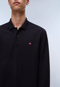 Camisa polo negra de manga larga con tejido texturizado, tapeta de tres botones y un pequeño logo de bandera bordado en el lado izquierdo del pecho.