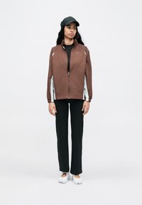 Καφέ μπουφάν με φερμουάρ και ανοιχτογκρί λεπτομέρειες, συνδυασμένο με μαύρο τοπ και μαύρο παντελόνι. Το μοντέλο φοράει μαύρο καπέλο και λευκά παπούτσια.