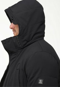 Juodas insulated striukė su aukštu apykakle ir gobtuvu. Geralus ant rankovės ir lygus, tekstūruotas audinys. Matomi užtrauktuko kišenės.