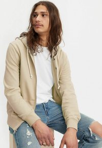 Beige Kapuzenpullover mit Reißverschluss und Kordeln, über einem weißen T-Shirt getragen. Hellblaue, zerrissene Jeans, auf einer Holzoberfläche sitzend. Langes, welliges Haar.