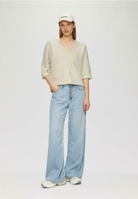 Beige, textuur V-hals top met driekwart mouwen, gecombineerd met lichtblauwe wijdvallende jeans en lichte sneakers. Pet met logo op het hoofd.