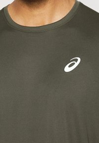 Tummanvihreä lyhythihainen urheilupaita, joka on valmistettu kevyestä, sileästä kankaasta; paidassa on vasemmalla rinnassa valkoinen logo ja pyöreä pääntie.