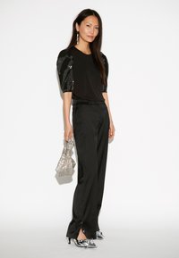 Schwarzes Oberteil mit Pailletten-Puffärmeln, schwarze hochtaillierte Hose, silberne Absätze und eine strukturierte silberne Umhängetasche. Glattes Material und taillierte Passform.