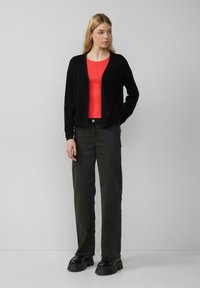 Cardigan noir sur un haut rouge, associé à un pantalon vert foncé et des chaussures noires imposantes. Tissu lisse, design ouvert à l'avant, manches longues.