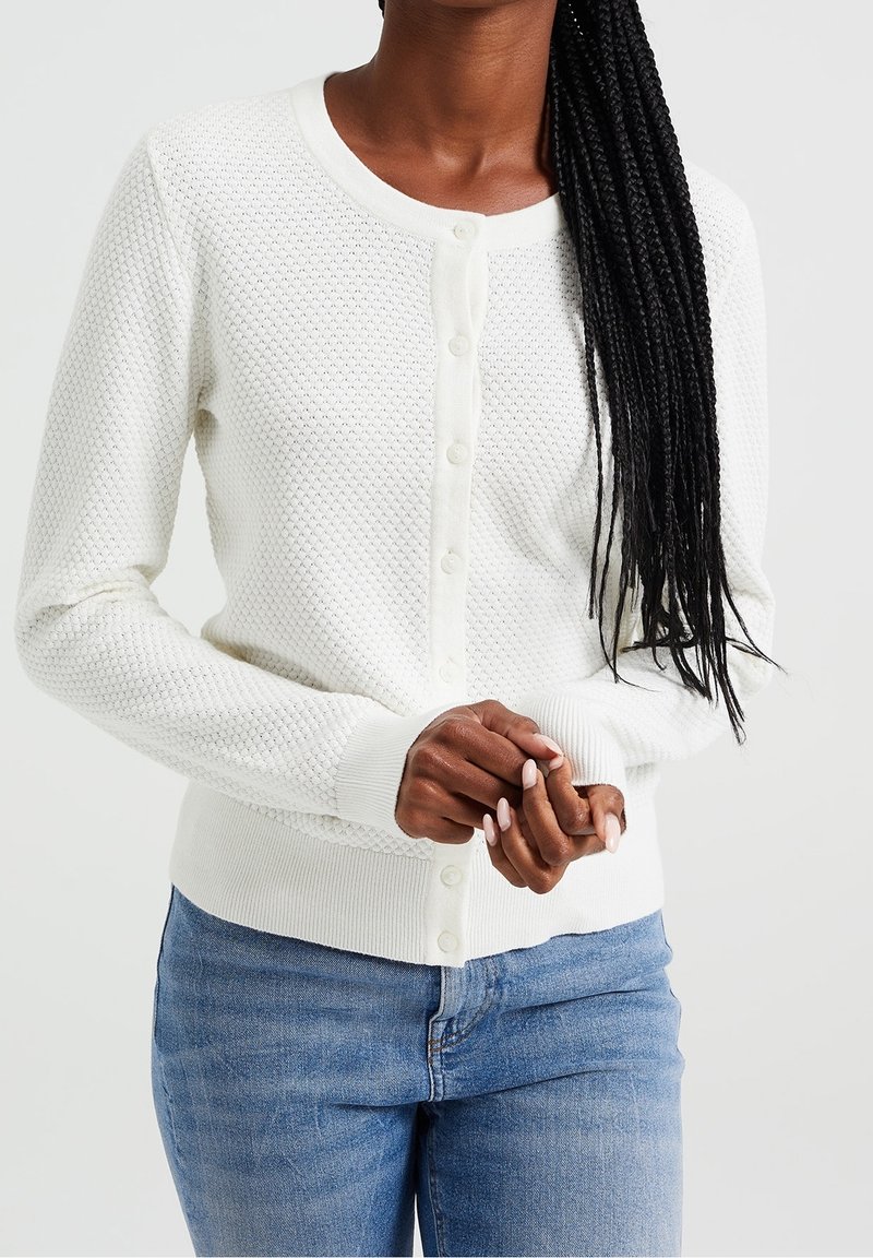 Cardigan blanc texturé avec un col rond, fermeture à boutons sur le devant, et bords-côtes aux poignets et à l'ourlet. Porté sur un jean bleu.