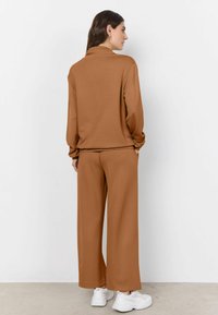 Hnedý, ľahký, dvojdielny outfit s golierom, dlhými rukávmi a voľnými širokými nohavicami. Nosený s bielymi teniskami.