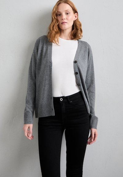 Grauer Cardigan mit durchgeknöpftem Front, gerippten Bündchen und lockerer Passform, getragen über einem weißen Hemd, kombiniert mit schwarzen hochtaillierten Jeans.