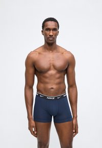 Muskuløs mann som står, iført marineblå Nike boxer shorts med svart midjebånd, ensfarget lys grå bakgrunn, nøytral uttrykk.
