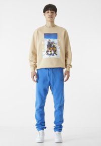 Beige genser med grafisk trykk, parret med blå sweatpants med snøring i livet og ribbestrikkede ankler. Modellen har på seg hvite joggesko.