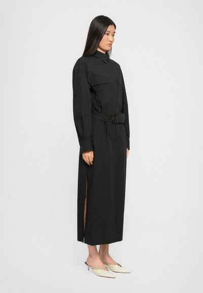 Woman wearing a long black belted shirt dress with side slits and beige pointed-toe kitten heel mules, standing against a plain white background.