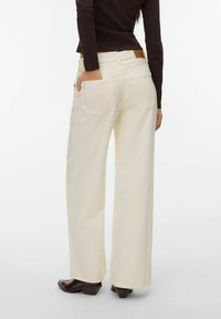 Personne portant un pantalon large crème et des bottes noires, main dans la poche arrière, sur fond blanc uni.