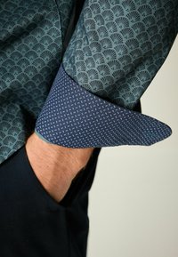 Hemd mit Knopfverschluss in einem türkisfarbenen Fächerdesign auf dem Ärmel und einem kontrastierenden dunkelblauen Manschette mit weißen Punkten. Glatte Stoffoberfläche.