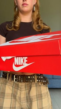 Caixa de sapatos vermelha da Nike com o logotipo branco, segurada por uma pessoa. A caixa tem um acabamento brilhante, forma retangular e uma tampa parcialmente aberta.