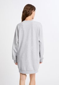 Donna con lunghi capelli castani che indossa un vestito felpa grigio chiaro a maniche lunghe e larghe, di spalle su uno sfondo bianco.