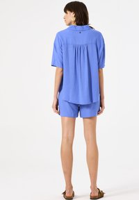 Camisa de lino azul con mangas cortas y espalda plisada, combinada con pantalones cortos a juego. Presenta un cuello y un detalle de botón redondo.