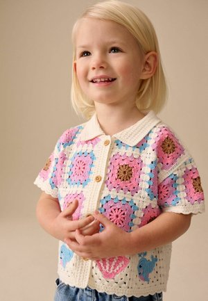 Ungt barn med blondt hår smiler iført en farverig hæklede skjorte med knapper, dekoreret med mønstre i pink, blå og brune farver samt havdyr.