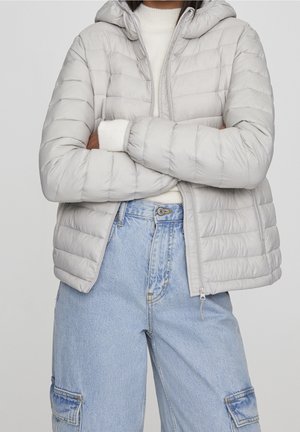 Personne portant une doudoune gris clair par-dessus un pull blanc, les bras croisés, associée à un pantalon cargo ample en denim bleu clair.