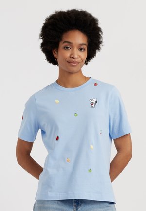 Femme aux cheveux courts et bouclés portant un t-shirt bleu clair avec de petits fruits brodés et le personnage Snoopy sur la poitrine.