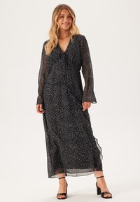 Robe longue à manches longues noire avec des pois blancs, silhouette fluide, en tissu léger et transparent. Elle présente un col en V et des manches évasées.