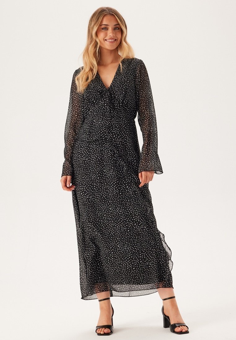 Robe longue à manches longues noire avec des pois blancs, silhouette fluide, en tissu léger et transparent. Elle présente un col en V et des manches évasées.