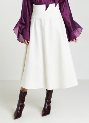 Donna che indossa una gonna midi bianca a vita alta, una blusa viola di seta con maniche svasate e stivaletti neri a punta di colore viola scuro.