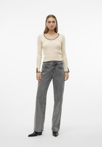 Maglia beige a maniche lunghe con collo rotondo e bordi neri, abbinata a jeans grigi in denim a vita alta e stivali neri con punta affilata.