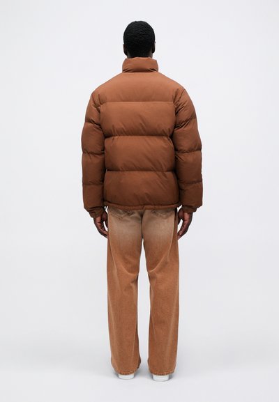 Brun pufferjakke med høj krave og quiltet design. Sammen med lysebrune, løstsiddende jeans og hvide sko.