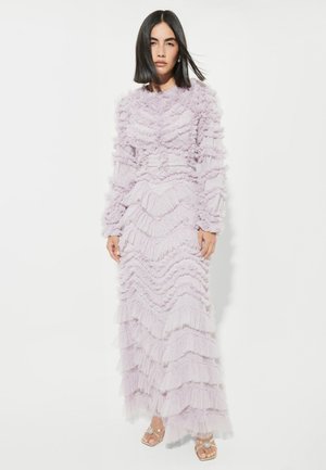 Mujer de pie con un vestido largo color lavanda con volantes, mangas largas y sandalias doradas abiertas en los dedos, sobre un fondo blanco.