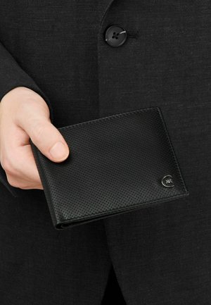 Käsi pitelee mustaa, teksturoitua nahkalompakkoa, jossa on pieni hopeinen logo, tumman napitetun pukutakin taustaa vasten.