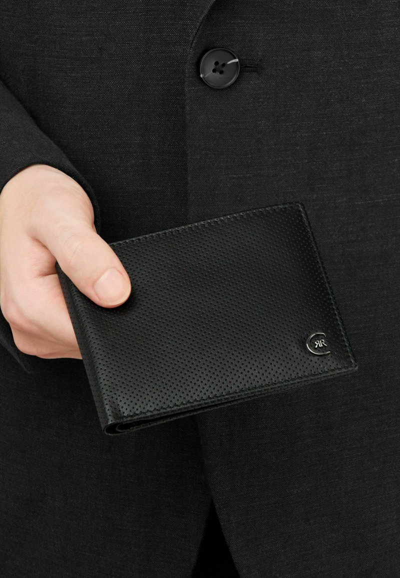 Mano sosteniendo una billetera de cuero negro texturizado con un pequeño logo plateado, sobre un fondo de chaqueta de traje oscura con botones.