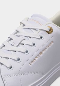 Λευκό αθλητικό παπούτσι Tommy Hilfiger με λευκά κορδόνια, χρυσές μεταλλικές οπές και χρυσό λογότυπο της μάρκας τυπωμένο στο λείο δερμάτινο επάνω μέρος.