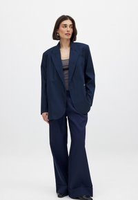 Blazer de gran tamaño en azul marino con cierre de un solo botón, amplios solapas y bolsillos con solapa, combinado con pantalones de pierna ancha a juego. Textura de tela suave.