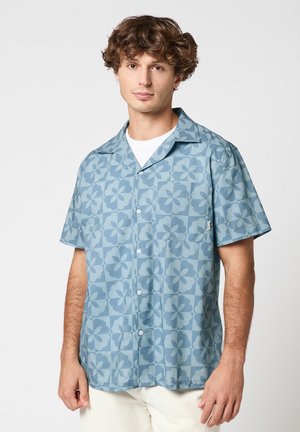 Jeune homme portant une chemise bleue à manches courtes avec un motif floral et un pantalon blanc, debout devant un fond uni.