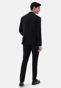 Schwarzer Anzug mit einer taillierten Silhouette, ausgestattet mit einem Schlitz hinten, kombiniert mit schwarzen Schuhen und einem weißen Hemd.