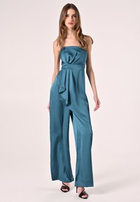 Tuta satiné teal senza spalline, con dettagli drappeggiati in vita, taglio a gamba larga e superficie liscia. Indossato con sandali neri con cinturini.