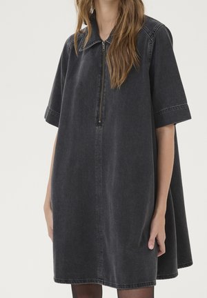 Frau trägt locker sitzendes schwarzes Denim-Kleid mit kurzen Ärmeln, vorderem Reißverschluss und Kragen vor schlichtem hellem Hintergrund.